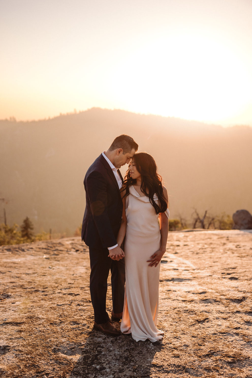 A couple dressed formally stands on rocky terrain, holding hands and looking at the mountain landscape during sunset.