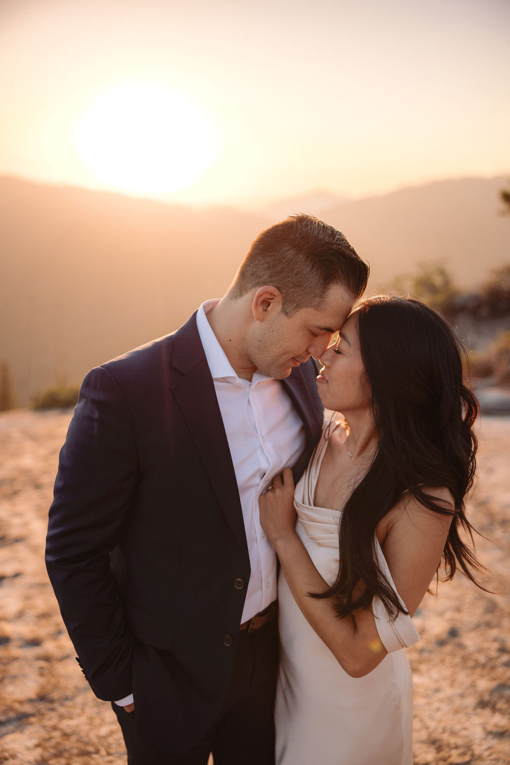 A couple dressed formally stands on rocky terrain, holding hands and looking at the mountain landscape during sunset.