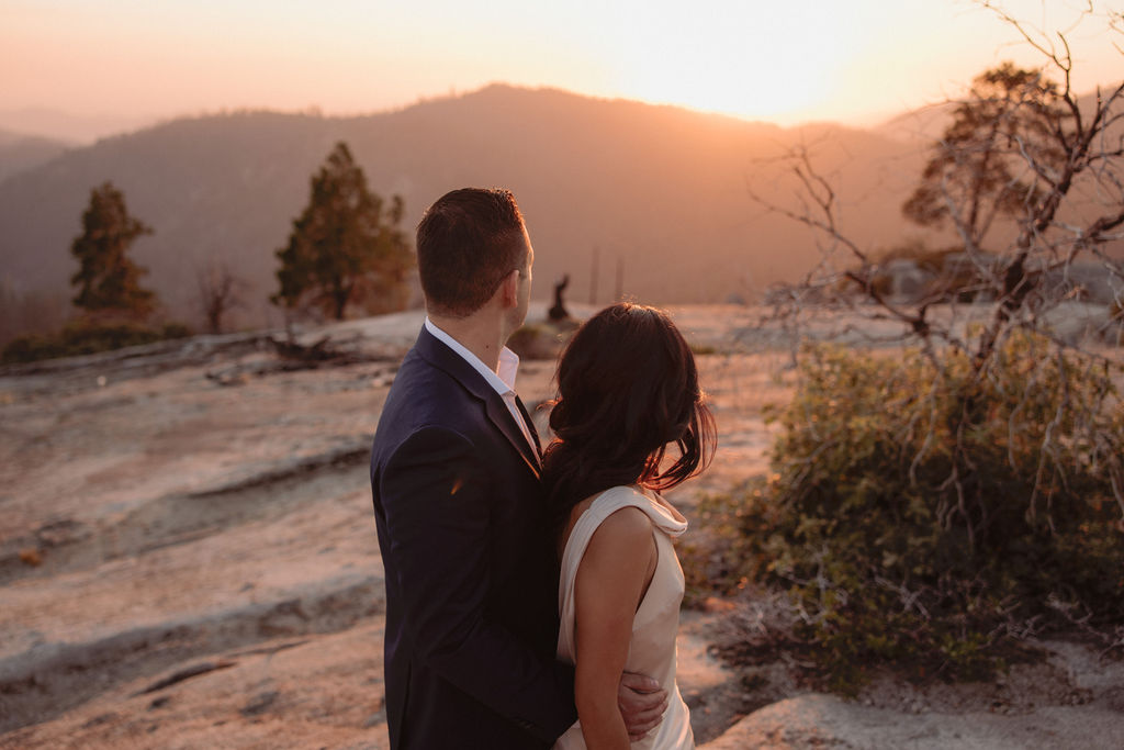 A couple dressed formally stands on rocky terrain, holding hands and looking at the mountain landscape during sunset.