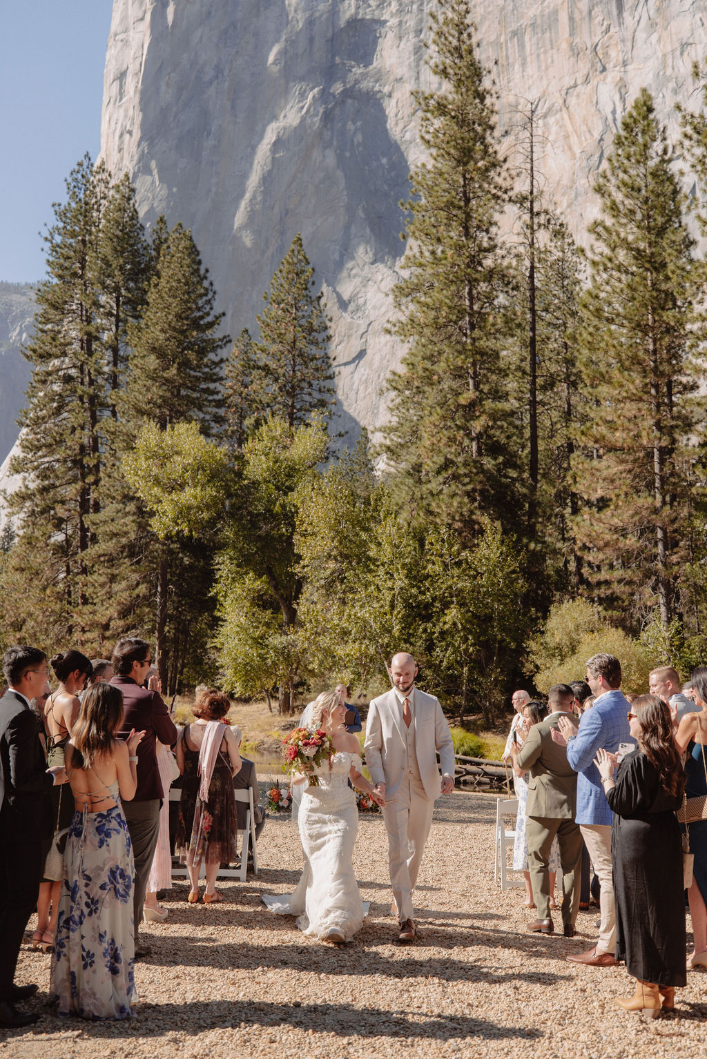 A wedding ceremony takes place outdoors with guests seated, surrounded by trees and mountains, with a large rock formation in the background.