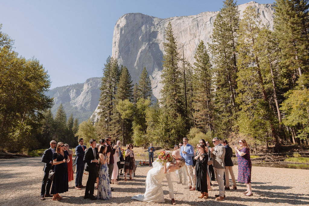 A bride and groom stand at an outdoor wedding ceremony with four attendants and an officiant by a river, surrounded by trees and mountains.