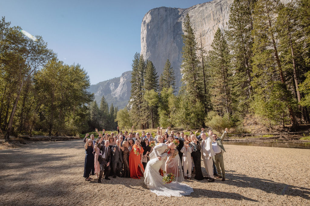 A bride and groom stand at an outdoor wedding ceremony with four attendants and an officiant by a river, surrounded by trees and mountains.