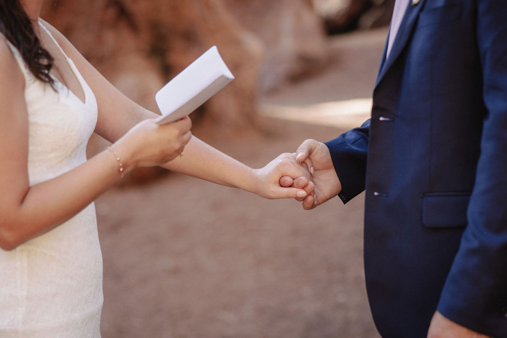 Bride in a white dress holds a piece of paper and reads vows while holding hands with a groom in a blue suit during an outdoor wedding ceremony.
