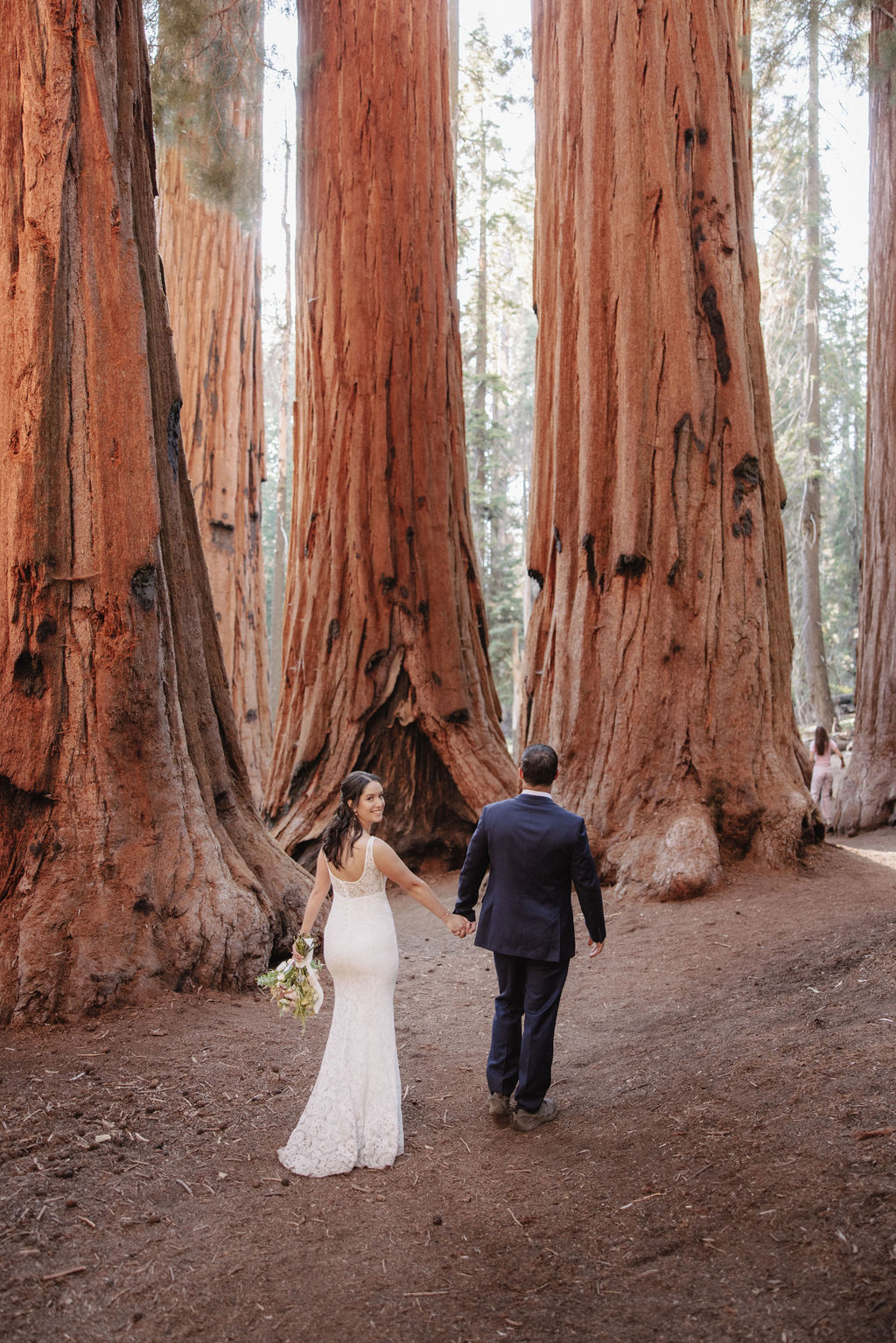 A bride and groom walk hand-in-hand through a forest of tall, reddish-barked trees, with sunlight filtering through the branches.