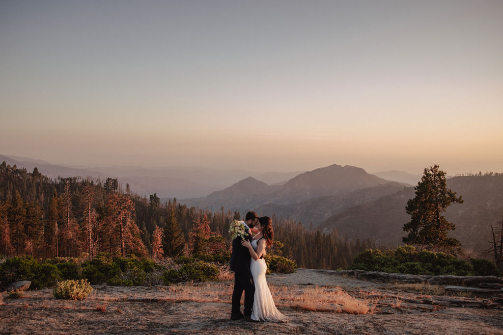 A bride and groom embrace on a rocky overlook at sunset with mountains and trees in the background.