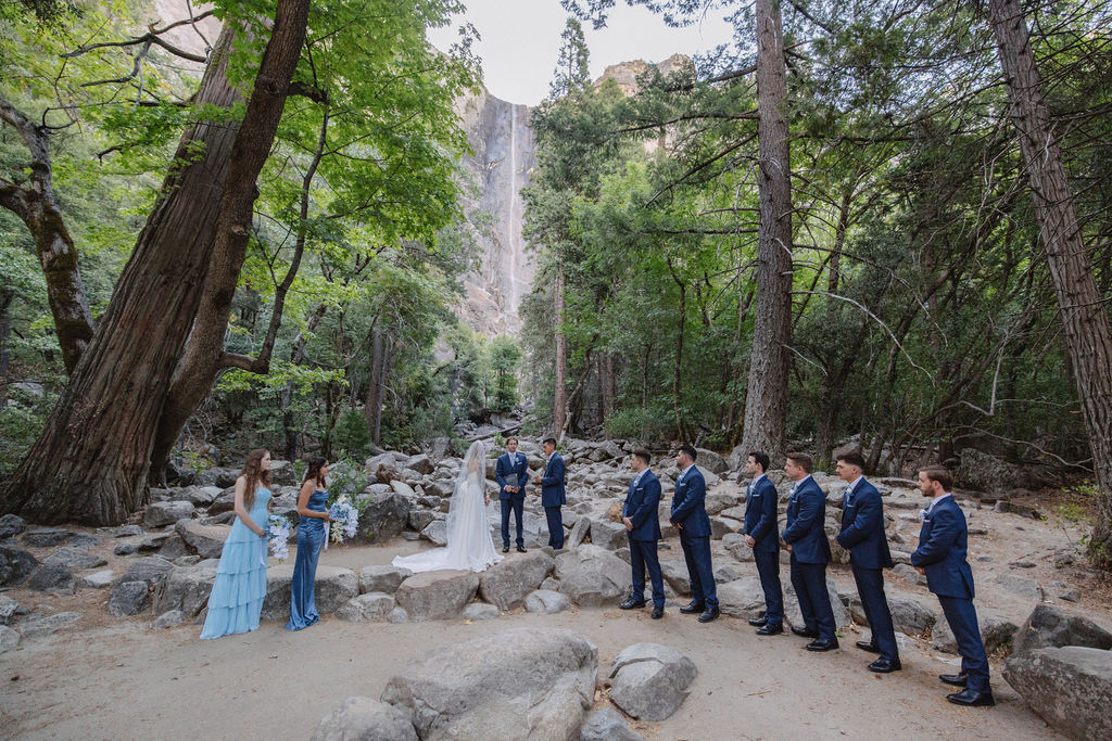 A wedding ceremony takes place outdoors on rocky ground, with a waterfall and tall trees in the background. The couple stands facing an officiant, with attendants nearby. Elopement vs. Micro Wedding: Which Is Right for You?