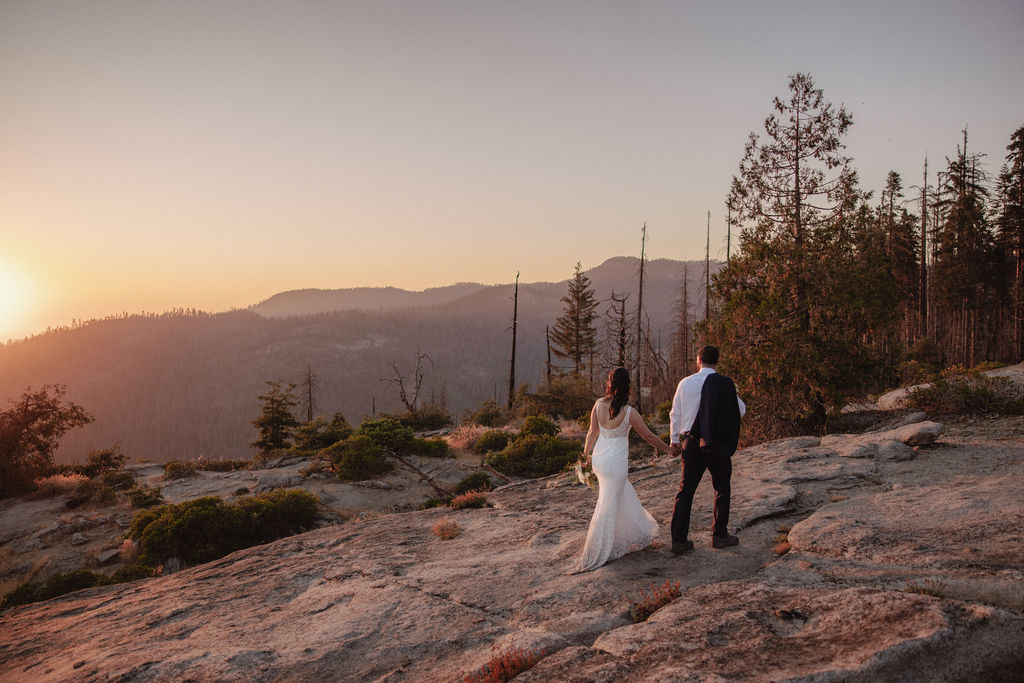 A bride and groom embrace on a rocky overlook at sunset with mountains and trees in the background.