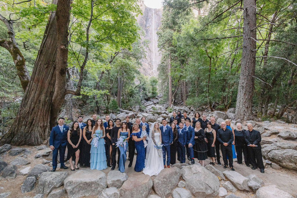 A wedding ceremony takes place outdoors on rocky ground, with a waterfall and tall trees in the background. The couple stands facing an officiant, with attendants nearby. Elopement vs. Micro Wedding: Which Is Right for You?