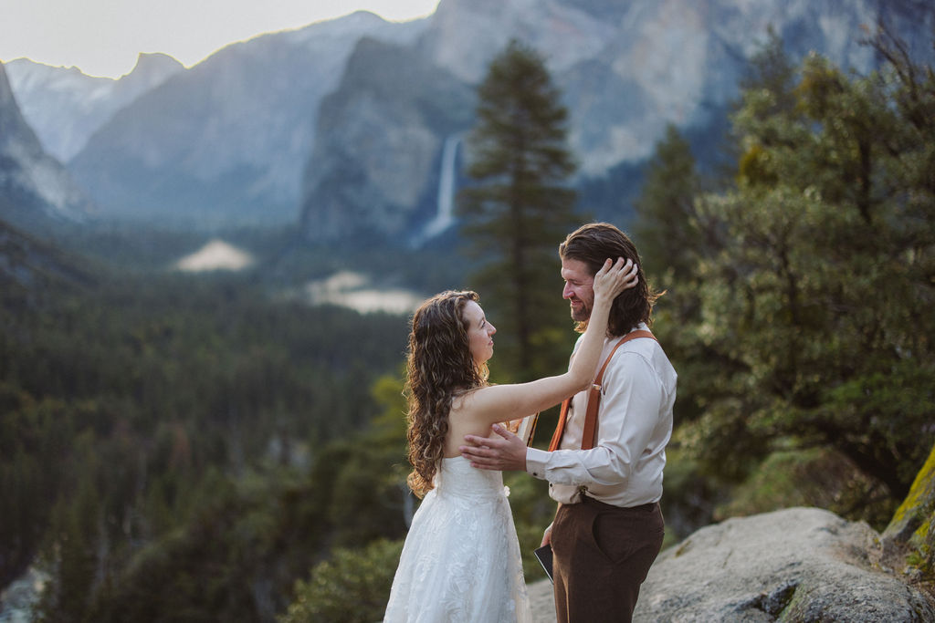 A bride and groom stand facing each other on a rocky overlook with mountains, trees, and a waterfall in the background.