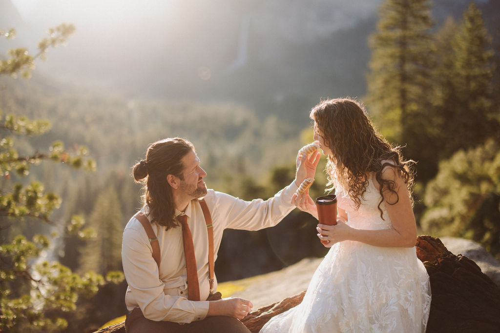 A man in a shirt and suspenders gently touches the face of a woman in a white dress while sitting outdoors on a sunlit forested hillside.