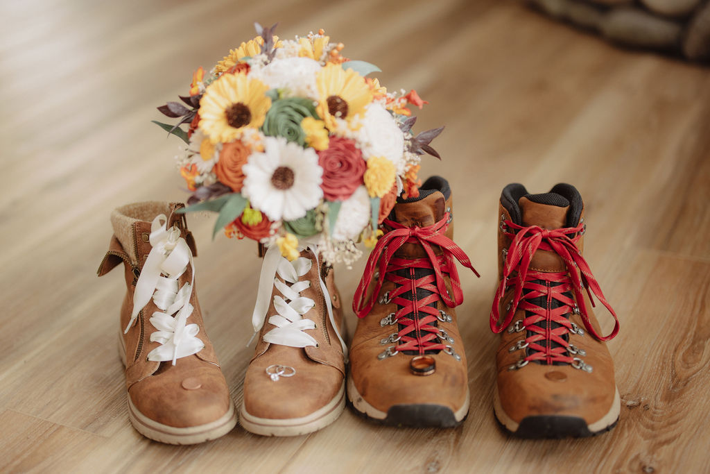 Two pairs of brown hiking boots with a bouquet of colorful flowers placed between them on a wooden floor.