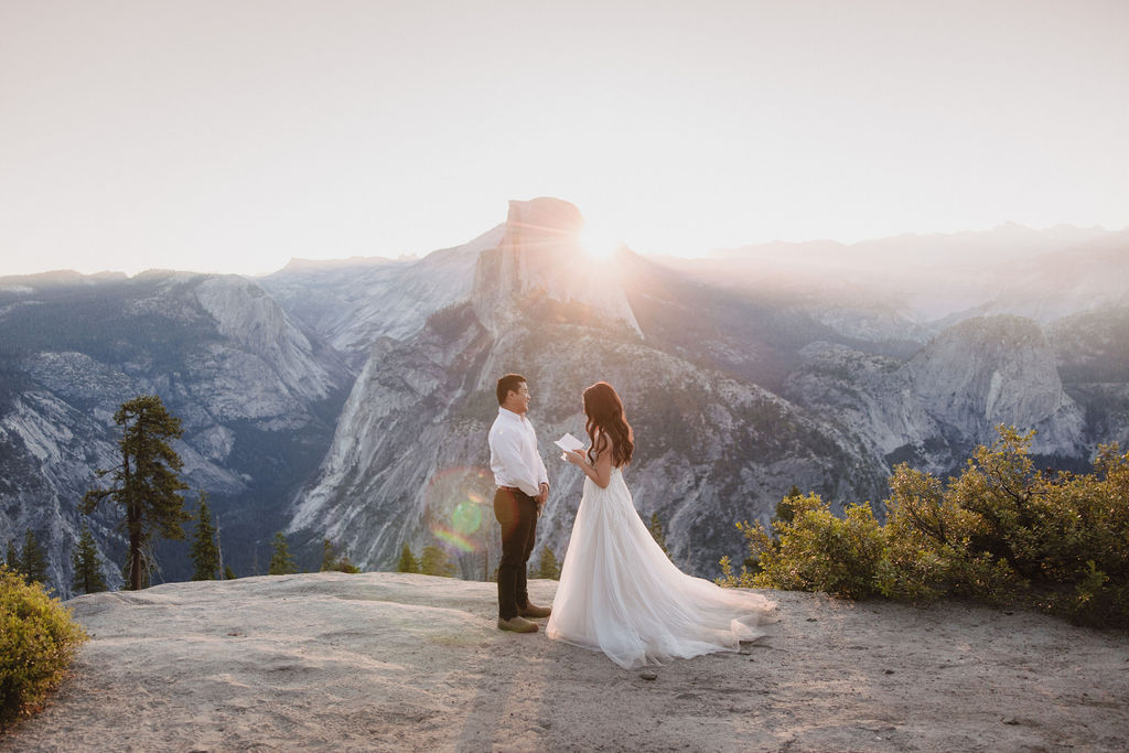 A bride and groom stand facing each other on a rocky ledge at sunset, with a mountain and sun rays in the background.