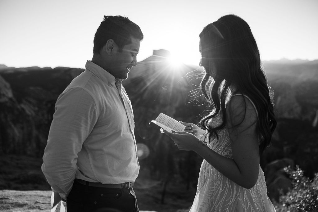 A bride and groom stand facing each other on a rocky ledge at sunset, with a mountain and sun rays in the background.