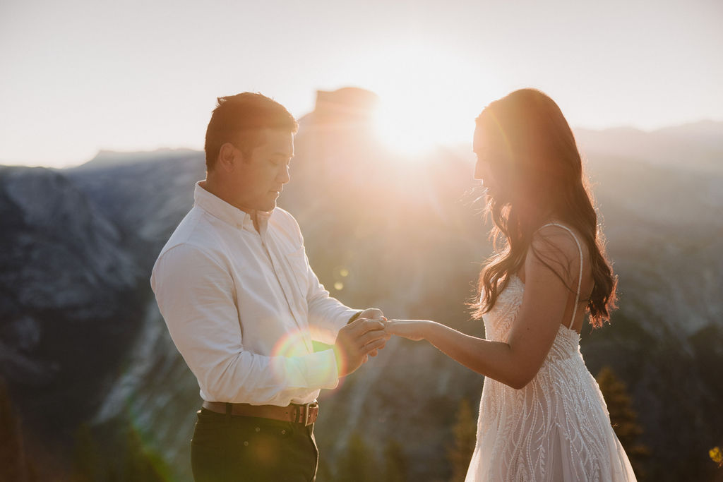 A bride and groom stand facing each other on a rocky ledge at sunset, with a mountain and sun rays in the background.
