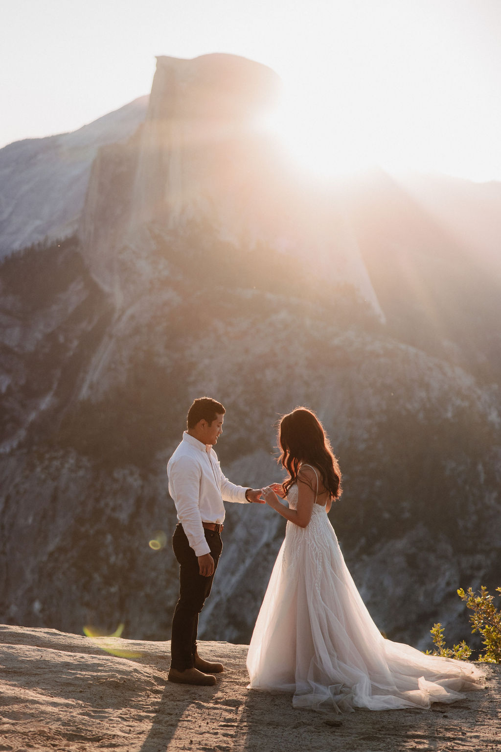 A bride and groom stand facing each other on a rocky ledge at sunset, with a mountain and sun rays in the background.