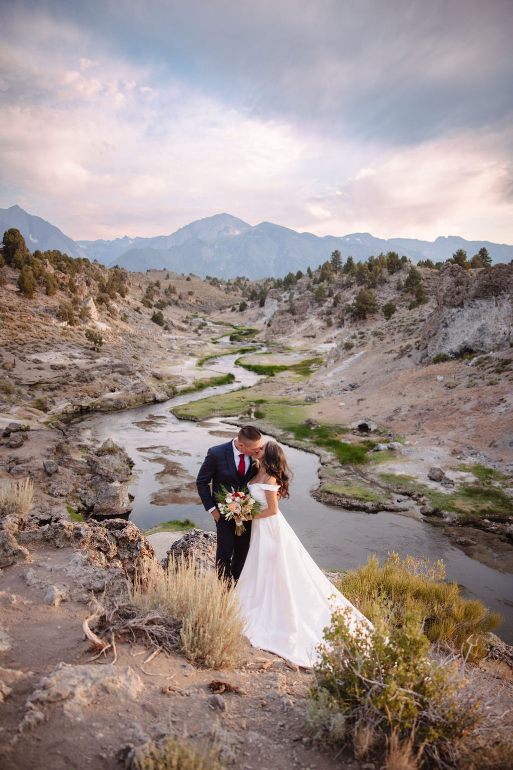 A couple dressed formally stands on rocky terrain, holding hands and looking at the mountain landscape during sunset.