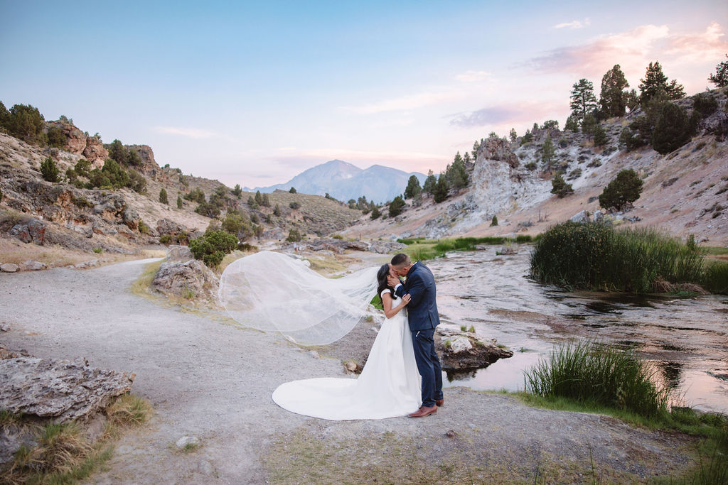 A couple dressed formally stands on rocky terrain, holding hands and looking at the mountain landscape during sunset.