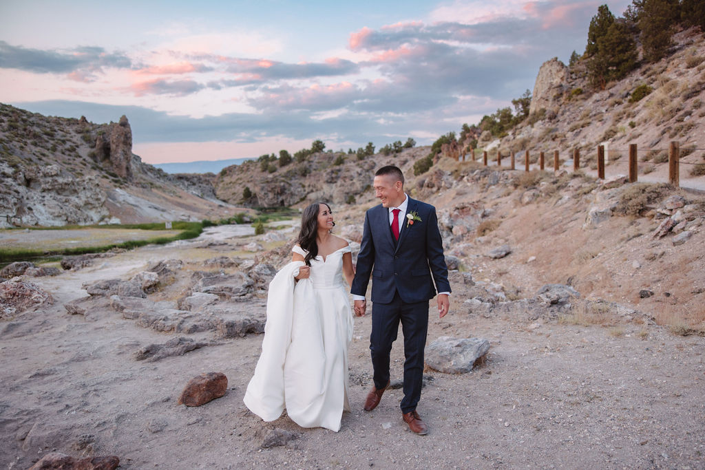 A couple dressed formally stands on rocky terrain, holding hands and looking at the mountain landscape during sunset.