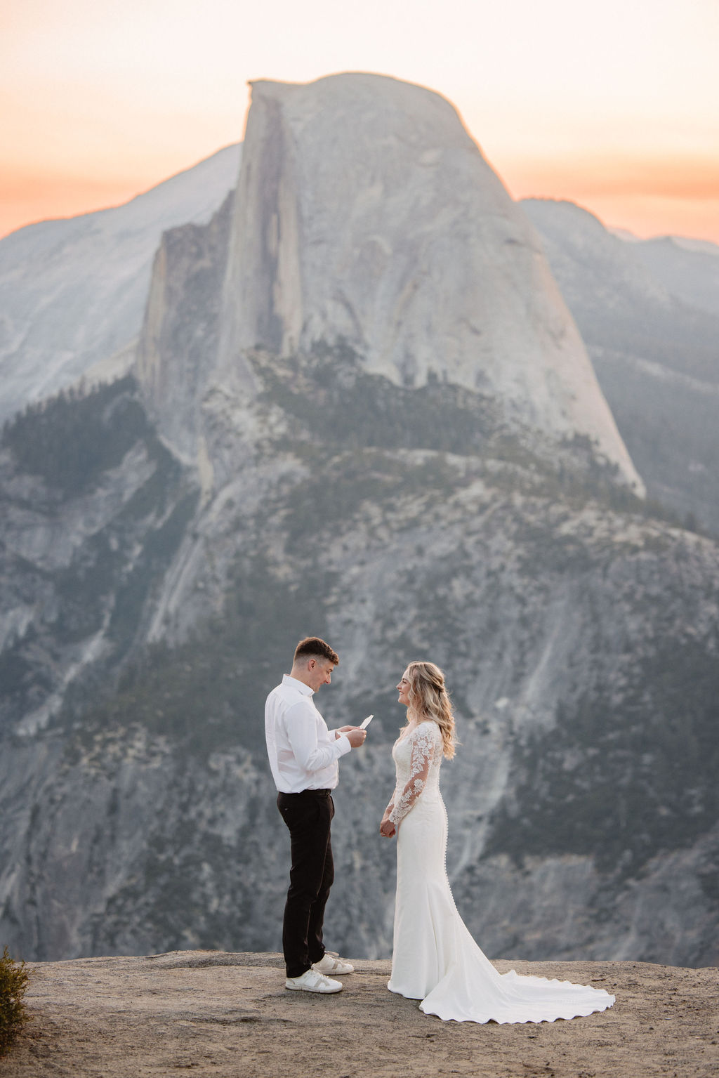 A bride and groom stand together on a rocky ledge with Half Dome and a mountainous landscape in the background at sunset as they exchange elopement vows