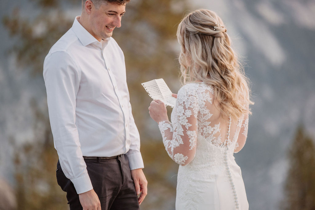 A bride and groom stand together on a rocky ledge with Half Dome and a mountainous landscape in the background at sunset as they exchange elopement vows