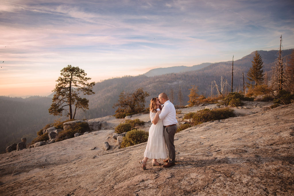 A couple stands close together outdoors, with mountains and trees in the background at sunset or sunrise.