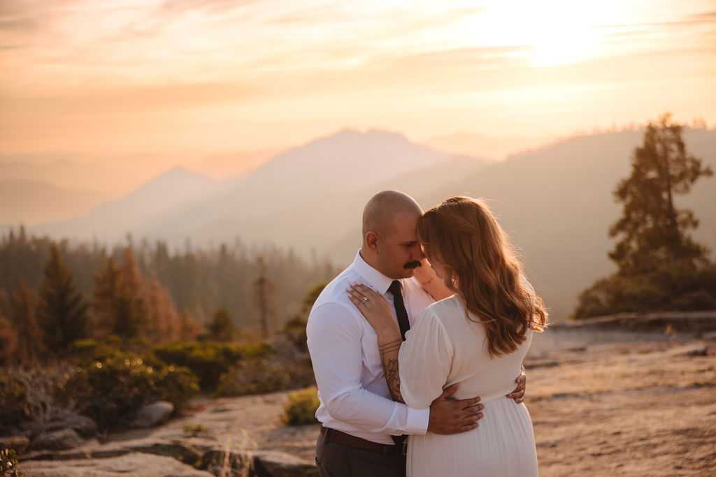 A couple stands close together outdoors, with mountains and trees in the background at sunset or sunrise.
