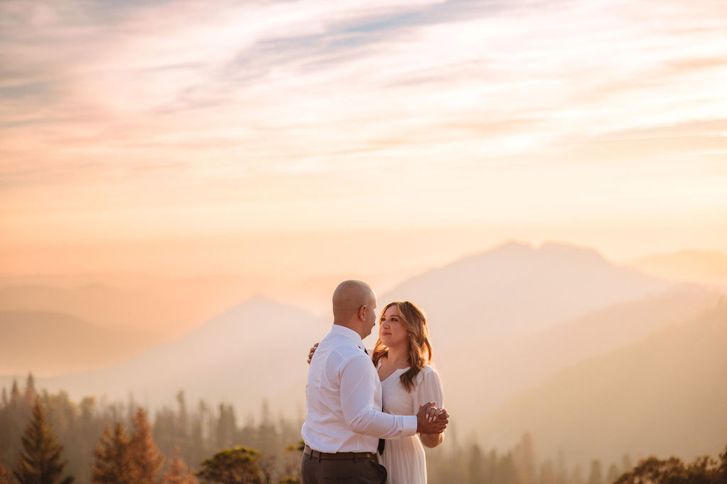 A couple stands close together outdoors, with mountains and trees in the background at sunset or sunrise.