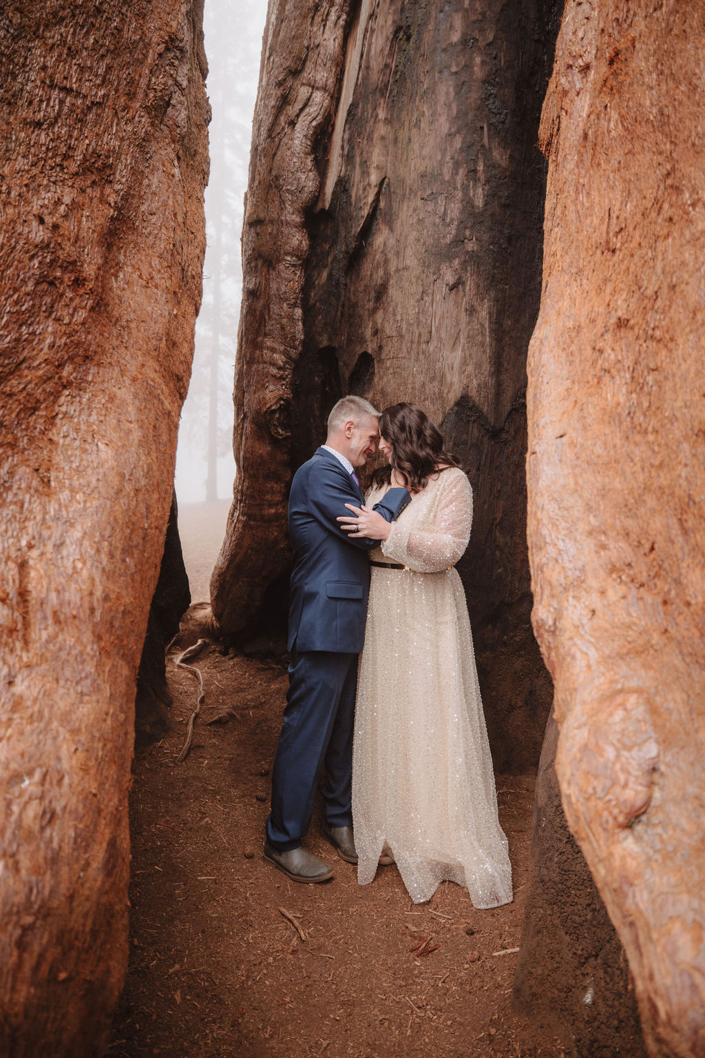 A couple stands close together, embracing between two large tree trunks in an outdoor setting.