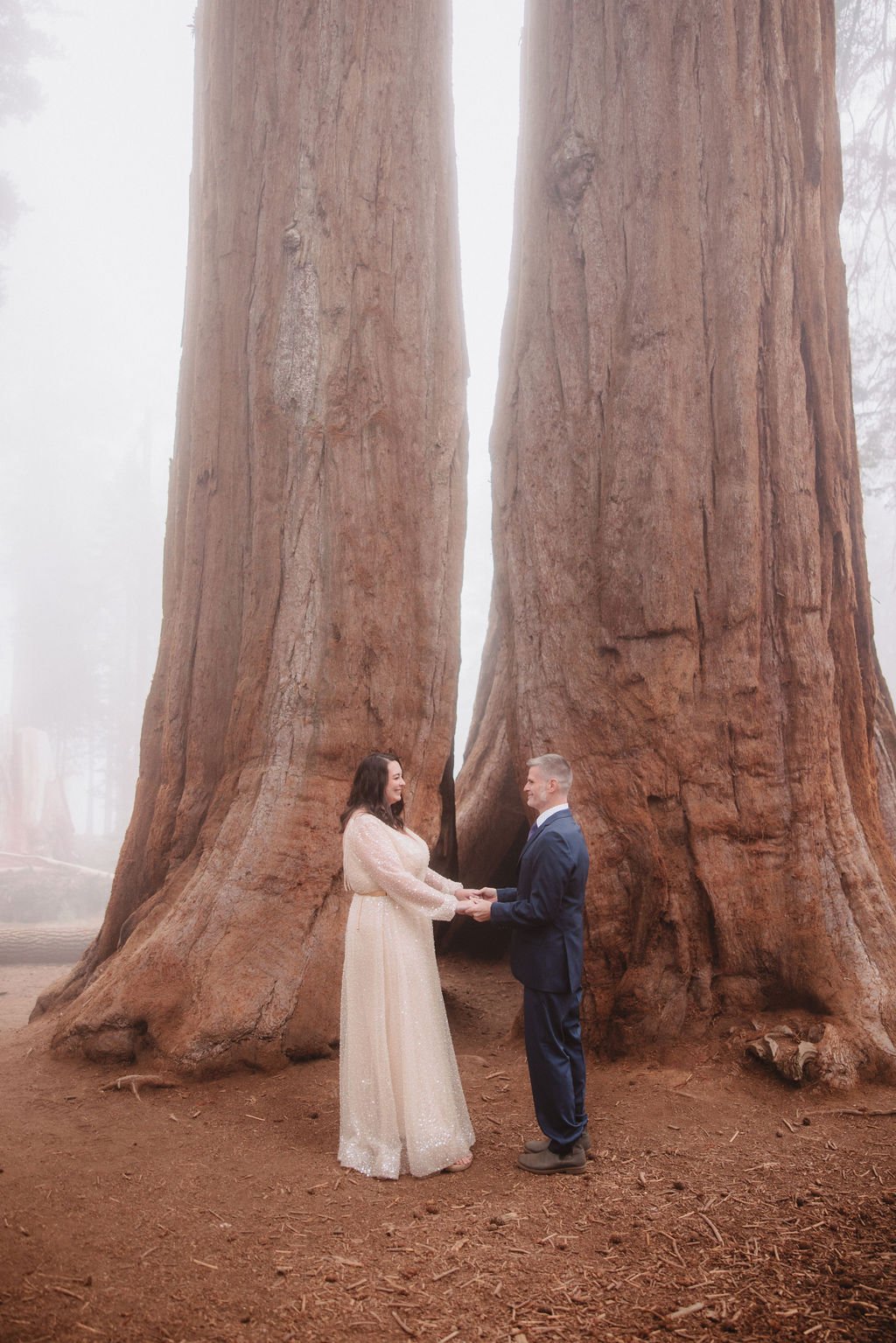 A couple stands close together, embracing between two large tree trunks in an outdoor setting.