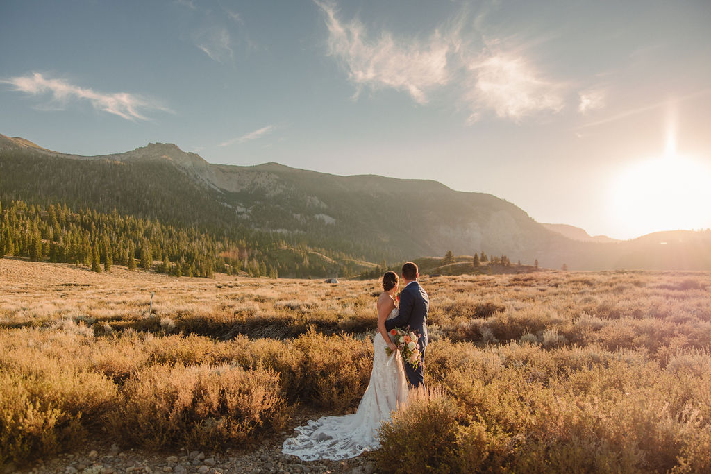 A bride and groom stand together in a sunlit field with mountains in the background during sunset.