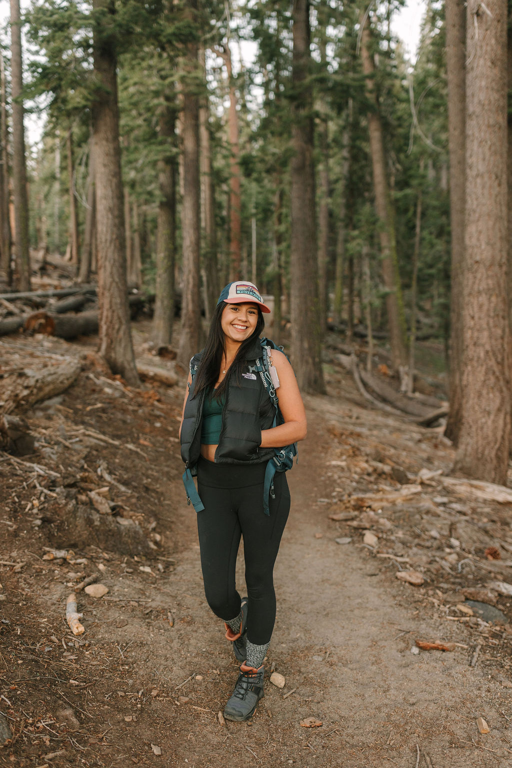 A person wearing a cap, vest, and athletic clothing walks on a dirt trail through a forest with tall trees. | Alyssa Michele Photo