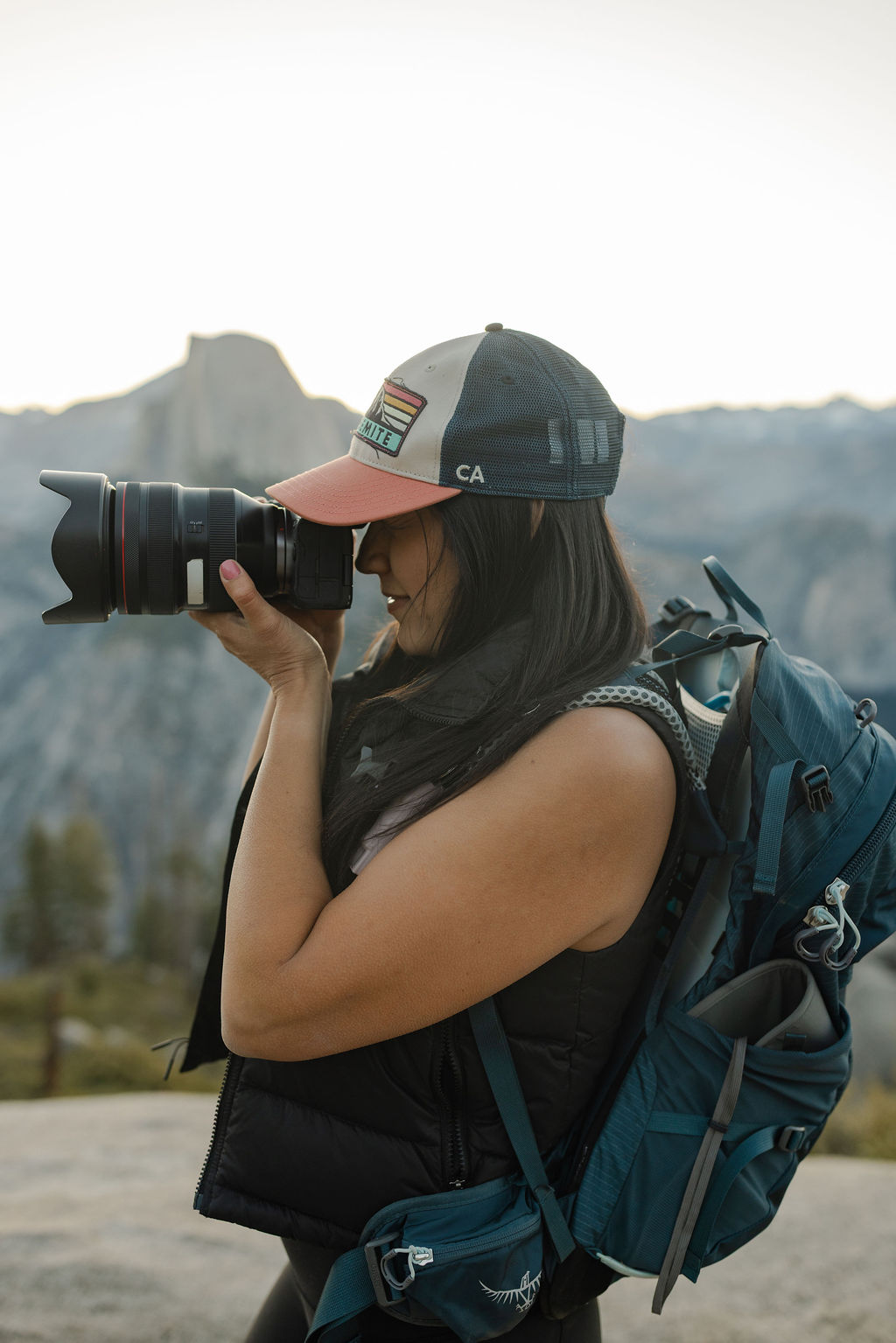 A woman wearing a cap and backpack takes a photo with a camera outdoors in a mountainous area.