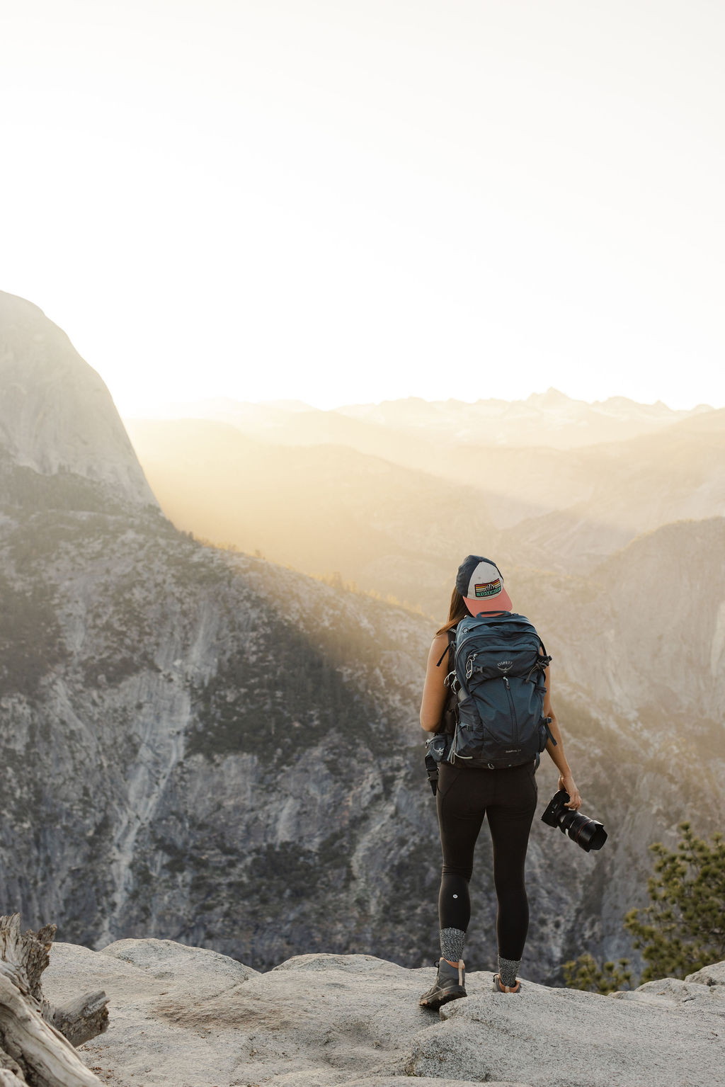 A woman wearing a cap and backpack takes a photo with a camera outdoors in a mountainous area.