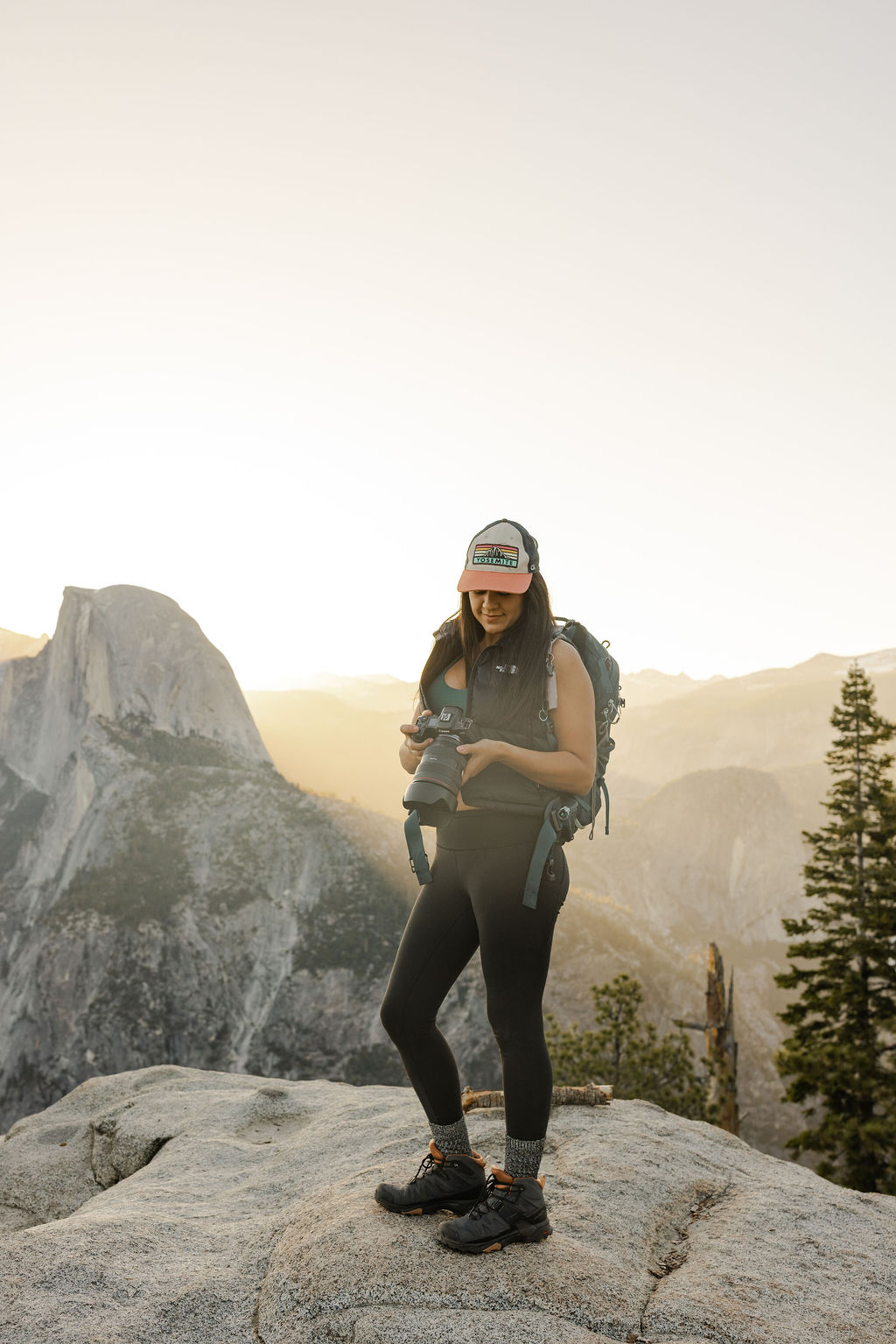 A woman wearing a cap and backpack takes a photo with a camera outdoors in a mountainous area.