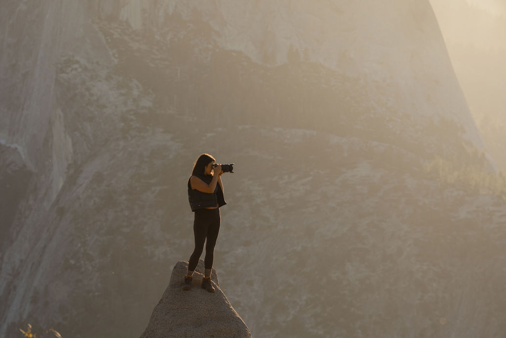 A woman wearing a cap and backpack takes a photo with a camera outdoors in a mountainous area.