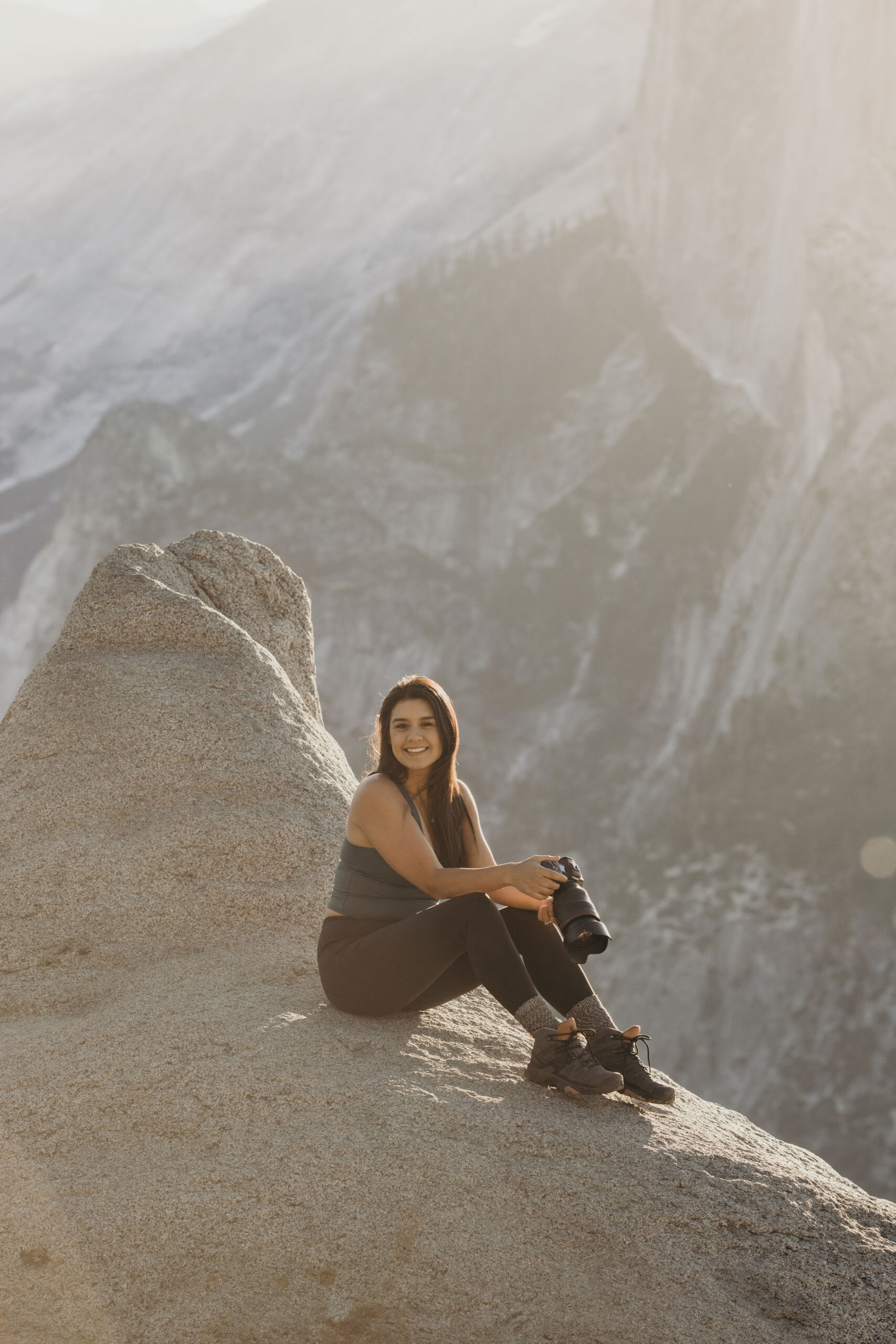 A person stands on a rocky ledge holding a camera, looking out at a sunlit mountain landscape in the background.