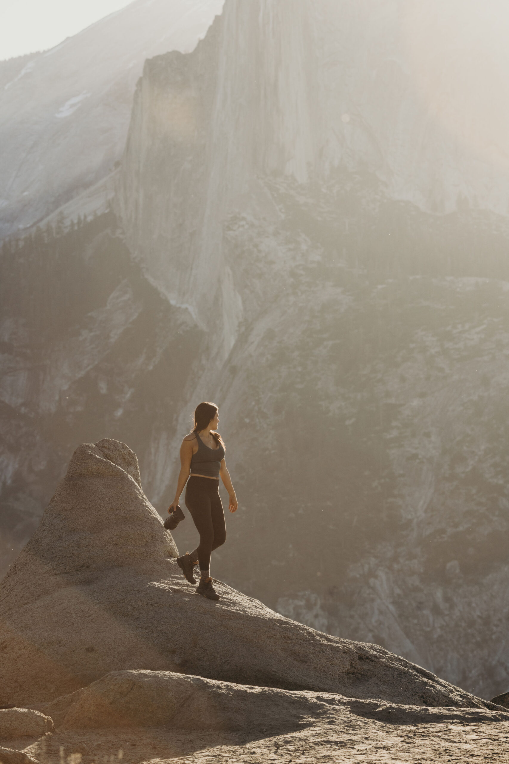 A person stands on a rocky ledge holding a camera, looking out at a sunlit mountain landscape in the background.