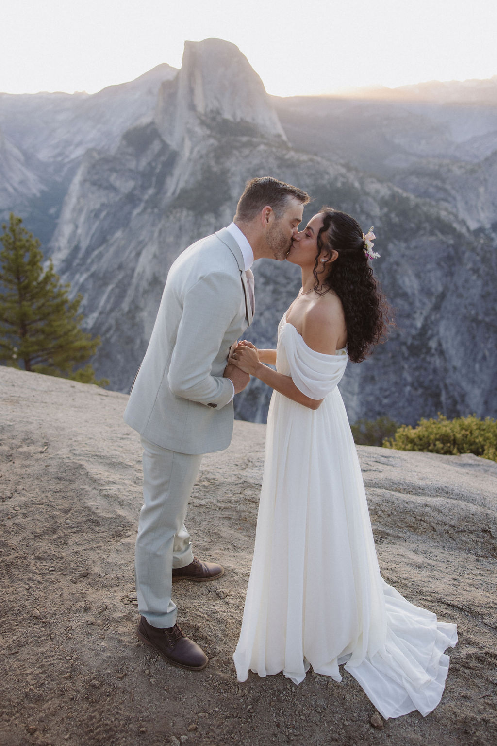 A bride and groom kiss outdoors on a rocky ledge with a mountainous landscape in the background.