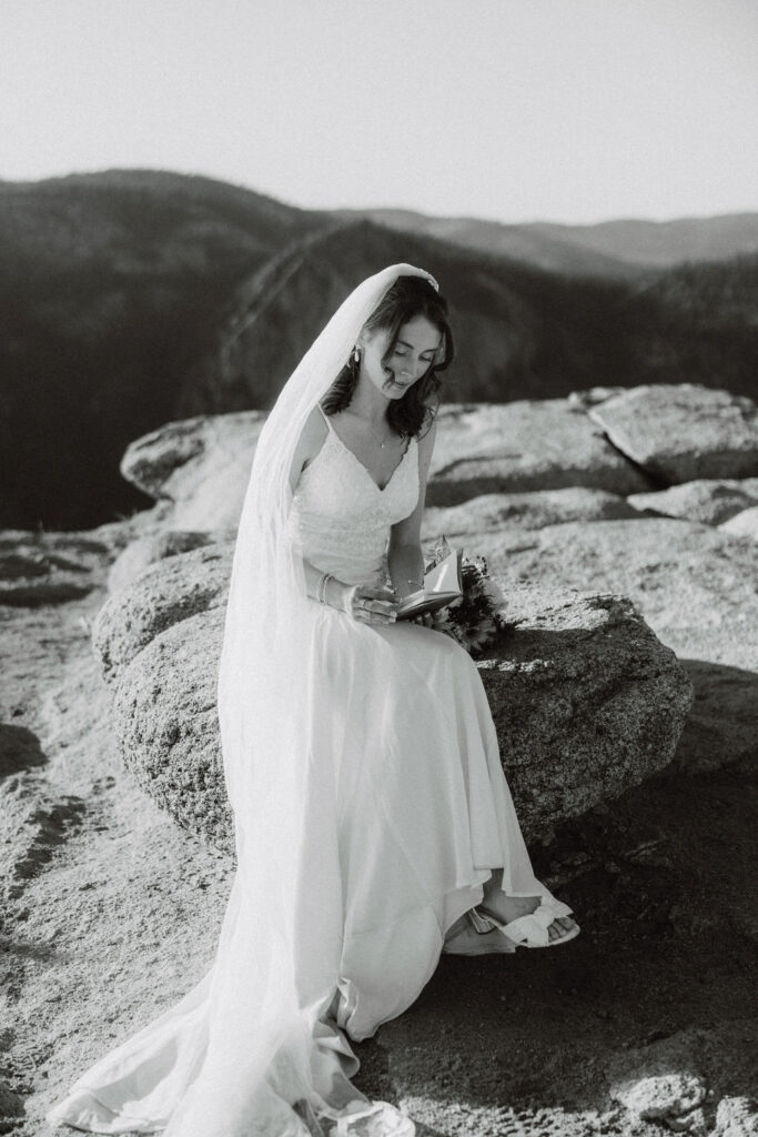 A bride in a wedding dress and veil sits on a rock outdoors, writing in a notebook, with mountains visible in the background taken by an experienced elopement photographer