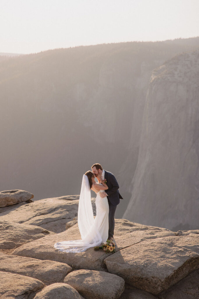 A bride and groom kiss on a rocky cliff with a mountainous background, dressed in formal wedding attire, under soft natural light by an experienced elopement photographer