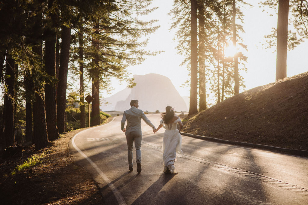 A couple holding hands walks down a tree-lined road at sunset, with a mountain visible in the background during their elopement adventure