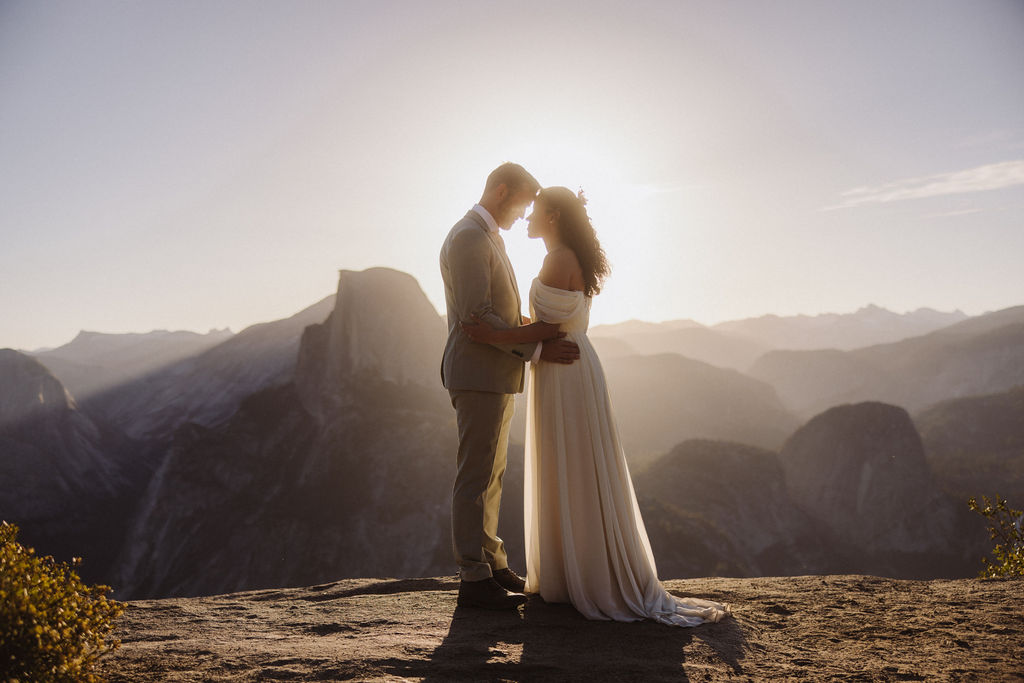 A bride and groom stand on a rocky mountaintop at sunset, sharing an intimate moment with mountains and sunlight in the background for their elopement adventure