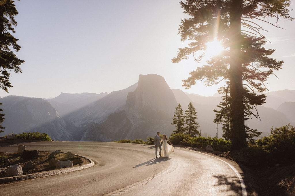 A couple holding hands walks down a tree-lined road at sunset, with a mountain visible in the background.