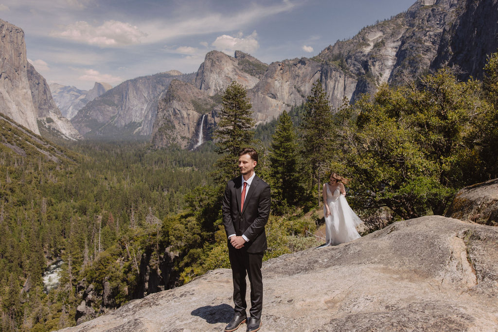 A groom in a suit stands on a rocky ledge with trees and mountains in the background, while a bride in a white dress approaches from behind.