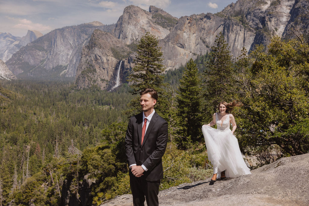 A groom in a suit stands on a rocky ledge with trees and mountains in the background, while a bride in a white dress approaches from behind.