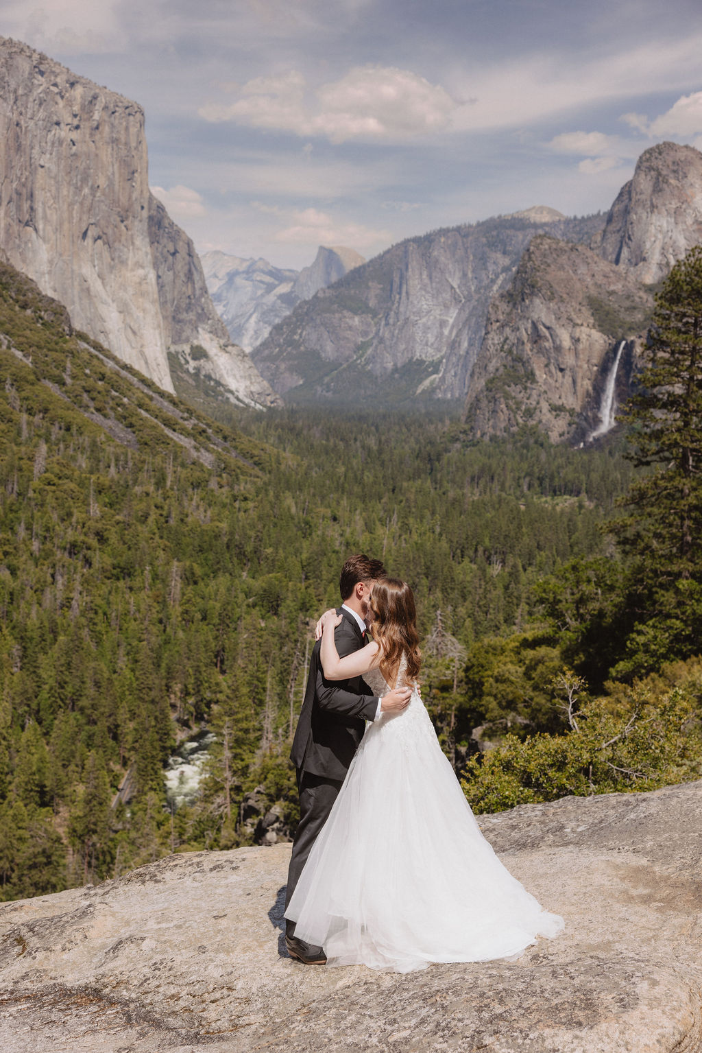 A bride and groom in wedding attire stand on a rocky overlook, embracing while facing a scenic view of mountains, cliffs, and forest in Yosemite National Park.