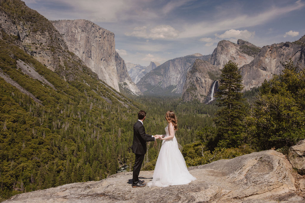 A bride and groom in wedding attire stand on a rocky overlook, embracing while facing a scenic view of mountains, cliffs, and forest in Yosemite National Park.