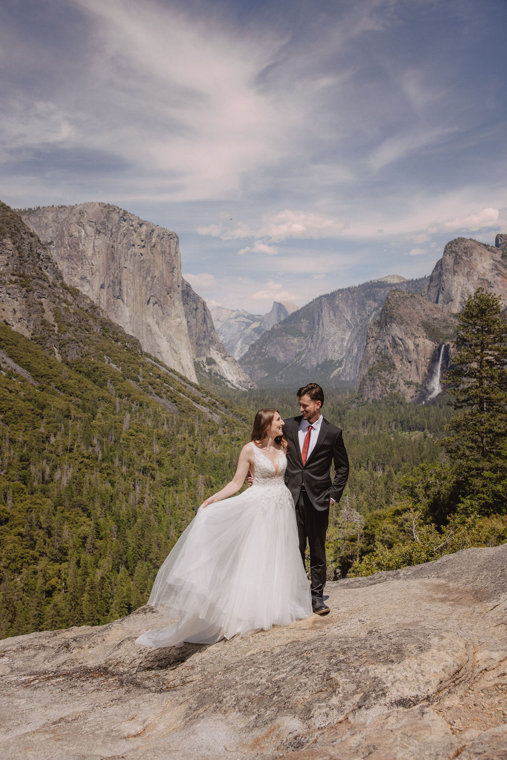 A bride and groom in wedding attire stand on a rocky overlook, embracing while facing a scenic view of mountains, cliffs, and forest in Yosemite National Park.