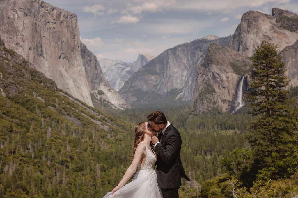 A bride and groom in wedding attire stand on a rocky overlook, embracing while facing a scenic view of mountains, cliffs, and forest in Yosemite National Park.