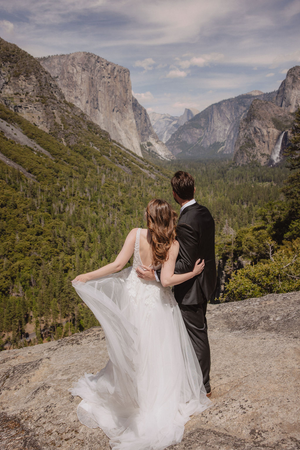 A bride and groom in wedding attire stand on a rocky overlook, embracing while facing a scenic view of mountains, cliffs, and forest in Yosemite National Park.